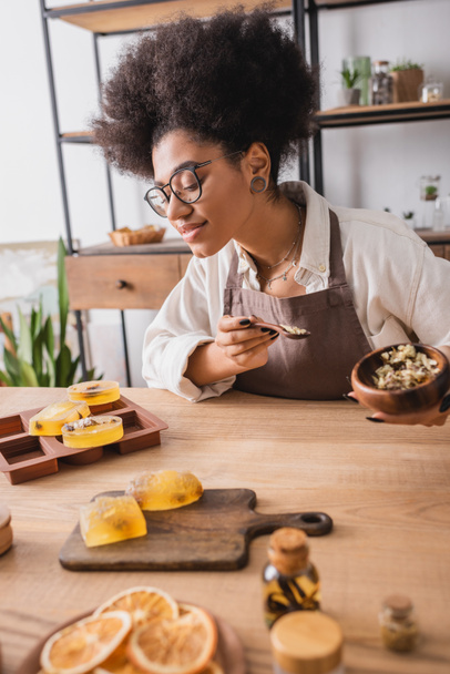 pleased african american craftswoman in eyeglasses holding dried herbs near soap bars and silicone mold on wooden table - Foto, imagen