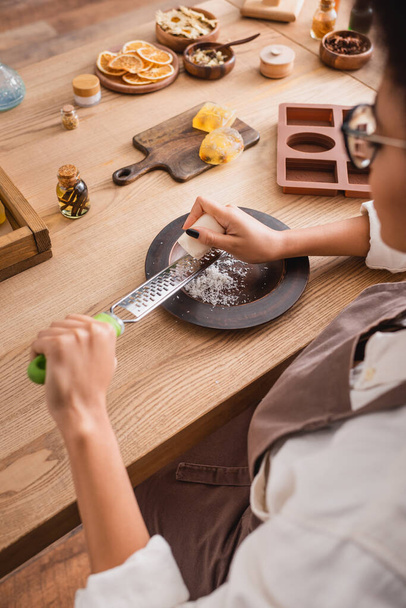 vista recortada de borrosa africana americana artesana rejilla jabón cerca de molde de silicona e ingredientes naturales en mesa de madera - Foto, imagen