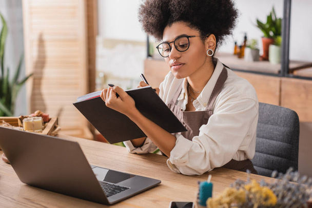 african american craftswoman in apron and eyeglasses writing in notebook near laptop and handmade products on blurred foreground - Photo, image