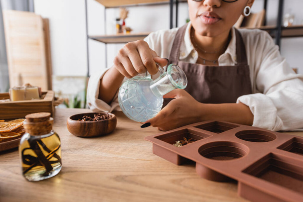 vista parcial de la mujer afroamericana sosteniendo jarra con jabón líquido cerca de molde de silicona e ingredientes naturales en taller de artesanía - Foto, imagen