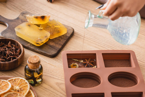partial view of african american craftswoman pouring liquid soap in silicone mold with dried herbs near essential oil and orange slices - Foto, afbeelding