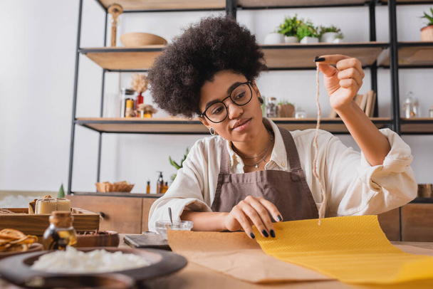 curly african american woman in eyeglasses and apron holding candle wick near wax sheet and natural ingredients on blurred foreground - Foto, imagen