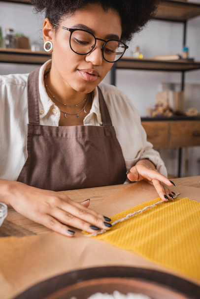artigiana afroamericana in occhiali e grembiule tenuta candela stoppino vicino foglio di cera su carta artigianale - Foto, immagini
