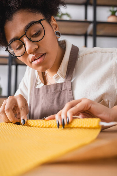 african american woman in apron and eyeglasses rolling wax sheet while making natural candle in craft workshop - Valokuva, kuva