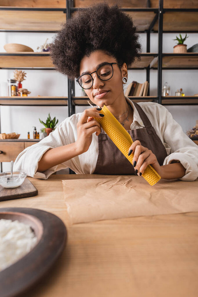 artesana afroamericana rizada en delantal y anteojos haciendo mango natural cerca de pergamino en mesa en taller - Foto, Imagen