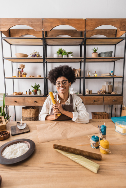 羊皮紙の近くに巻きワックスシートを持っている幸せなアフリカ系アメリカ人女性クラフトワークショップでテーブルの上に手作りキャンドル - 写真・画像