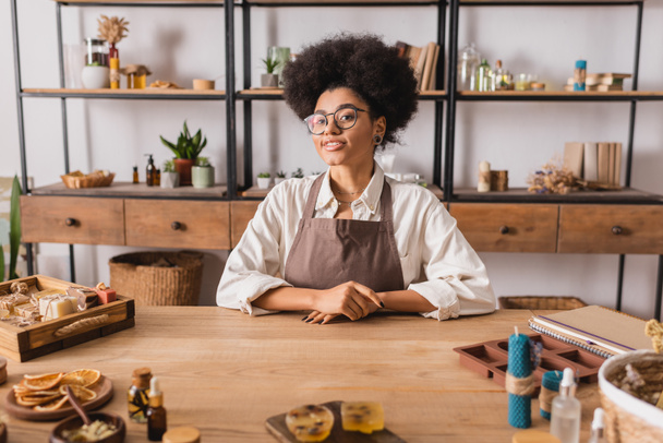 happy african american craftswoman in eyeglasses and apron looking at camera near natural products and ingredients in workshop - Photo, Image