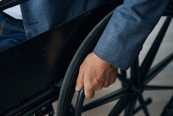 Zbliżenie zdjęcie męskiej dłoni na kole wózka inwalidzkiego, mężczyzny w szarej kurtce i dżinsach - Zdjęcie, obraz