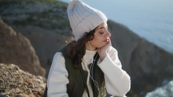 Smiling girl listening earphones music on mountain top. Relaxing serene woman enjoying song closing eyes in warm sunlight at ocean cliff view. Happy tourist taking break. Harmony stress free lifestyle - 映像、動画