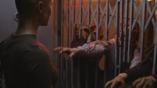 Eingesperrte Militärfrau mit rasiertem Kopf steht in dunklem, gruseligen Gebäude und blickt auf drei Zombies, die hinter dem Metalltor eines alten Fahrstuhls stecken und die Hände ausstrecken - Filmmaterial, Video