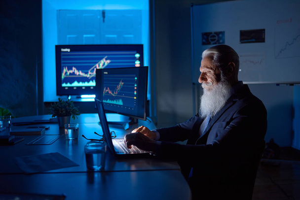 Вид сбоку пожилого мужчины-аналитика с седой бородой, работающего над нетбуком за столом с таблицами на мониторах в рабочем пространстве - Фото, изображение