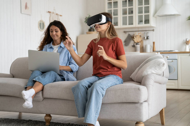 Concentrated little teen girl with VR glasses visits metaverse and plays modern video games with 3D personal presence effect. Enthusiastic schoolgirl is testing modern gadget sits on sofa near mother - Photo, Image