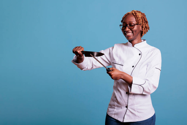 Mladá africká americká kuchařka s broušenými brýlemi a nožem na steak. Afro kuchař, kudrnaté vlasy, usmívající se, pomocí brousiče nožů. Restaurace pracovník udržuje kuchyňské nádobí. - Fotografie, Obrázek
