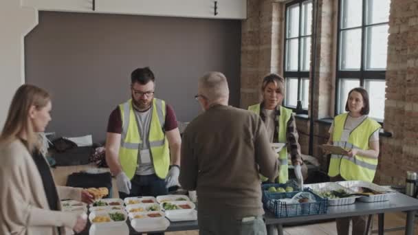 Medium long of diverse refugees getting disposable lunch boxes with hot meals and fresh green apples from young volunteers in food bank - Materiał filmowy, wideo