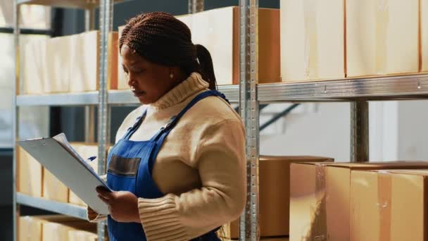 Young woman checking inventory list on clipboard, working with papers to see shipment order in storage room. African american person standing near shelves and racks in depot space. - Filmati, video
