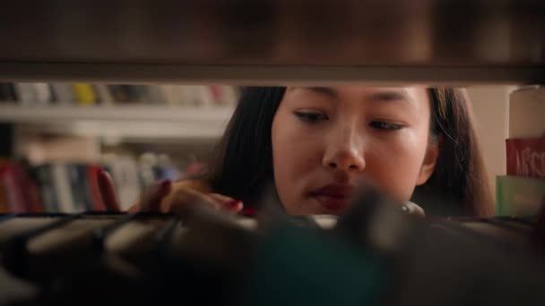 Close-up young asian woman browsing shelves books concentrated female student looking for necessary book to read on bookshelf searching right textbook to prepare for exam search literature in library - Imágenes, Vídeo