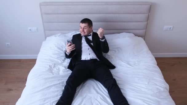 Office worker working lying on the bed at home, he is happy holding the phone in his hands. A happy successful man rejoices at the good news. You win the jackpot - Felvétel, videó