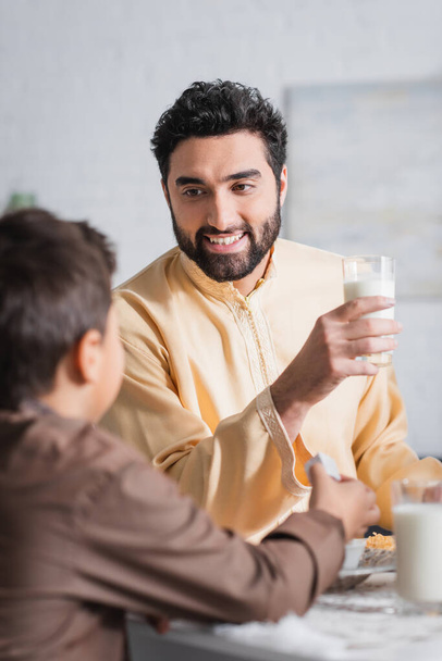 Smiling muslim father holding glass of milk and talking to blurred son during ramadan morning  - Valokuva, kuva