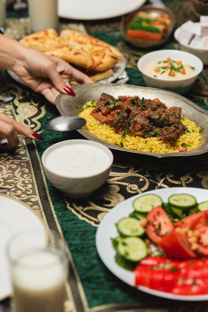 Vista cortada de mulher muçulmana colocando pilaf na mesa perto de iftar comida em casa  - Foto, Imagem