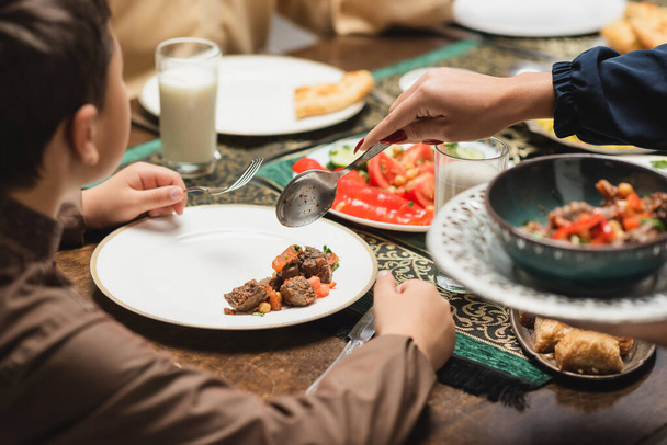 Μουσουλμάνα μητέρα σερβίρει φαγητό στο πιάτο κοντά στο γιο και ramadan δείπνο στο σπίτι  - Φωτογραφία, εικόνα