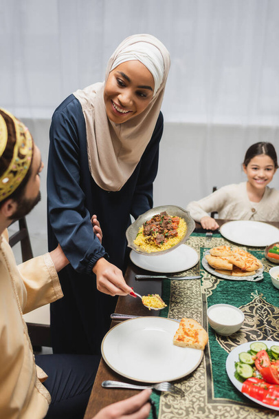 African american woman in hijab serving pilaf on plate near husband during ramadan at home  - Foto, Bild