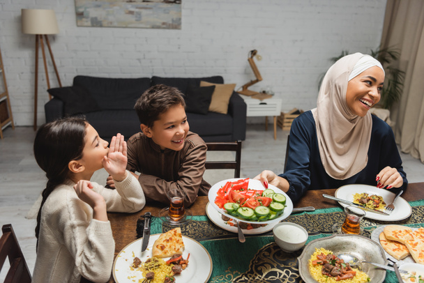 Веселые мусульманские дети разговаривают рядом с африканской американской мамой в хиджабе во время ифтара дома  - Фото, изображение