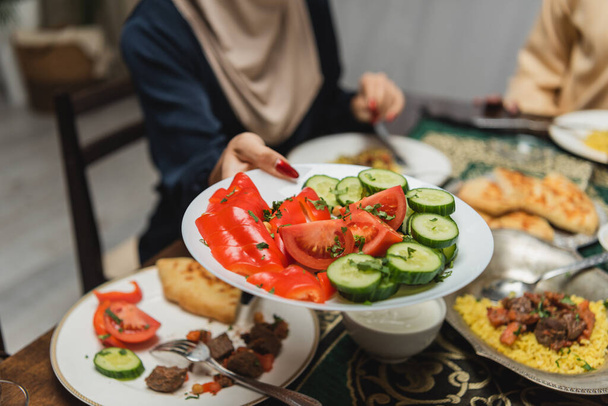 Обрезанный вид мусульманской женщины, держащей тарелку с овощами рядом с едой во время Ифтара дома  - Фото, изображение