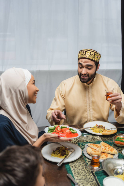 Χαμογελώντας μουσουλμάνος άνδρας κρατώντας το τσάι κοντά στη γυναίκα και το φαγητό κατά τη διάρκεια της iftar στο σπίτι  - Φωτογραφία, εικόνα