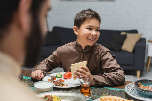Alegre chico musulmán mirando hacia otro lado cerca de la comida y papá durante iftar en casa  - Foto, Imagen