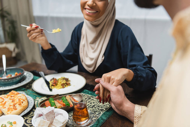 Καλλιεργημένη άποψη του μουσουλμανικού ζευγαριού που κρατιέται χέρι-χέρι κοντά στο φαγητό κατά τη διάρκεια του ιφτάρ στο σπίτι  - Φωτογραφία, εικόνα