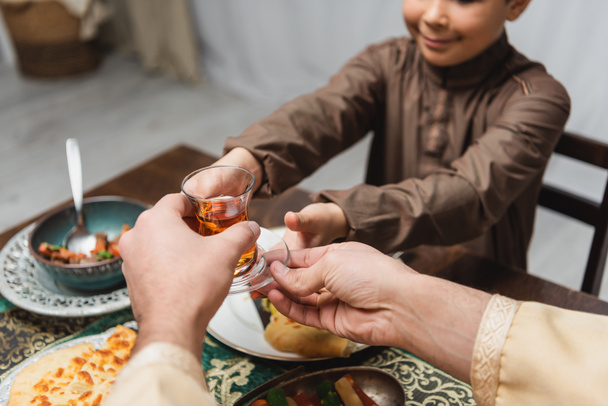 Ausgeschnittene Ansicht eines muslimischen Mannes, der seinem Sohn beim Iftar zu Hause ein türkisches Teeglas schenkt  - Foto, Bild