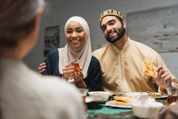Μουσουλμάνος άντρας αγκαλιάζει γυναίκα με μαντίλα κοντά σε θολή κόρη και φαγητό κατά τη διάρκεια του ραμαντάν στο σπίτι  - Φωτογραφία, εικόνα