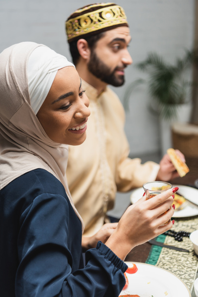 Femme afro-américaine souriante en hijab tenant un verre de thé près du mari flou pendant le ramadan à la maison  - Photo, image