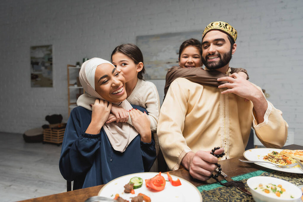 Niños musulmanes sonrientes abrazando a los padres durante la cena de ramadán en casa  - Foto, Imagen