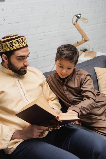 Muslim boy reading book with father on couch at home  - Фото, изображение