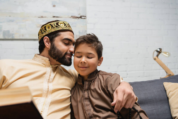 Χαμογελώντας μουσουλμάνος άνθρωπος με χάντρες προσευχή αγκαλιάζει γιο κοντά βιβλίο στον καναπέ στο σπίτι  - Φωτογραφία, εικόνα