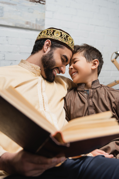 Cheerful middle eastern man and son reading blurred book at home  - Zdjęcie, obraz