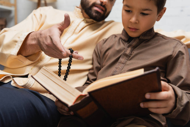 Moslim man met gebedskralen wijzend naar boek nabij zoon thuis  - Foto, afbeelding