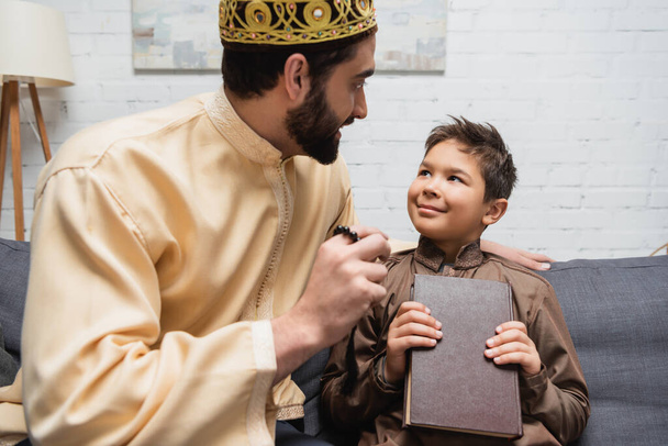 Padre musulmano che parla con il figlio che tiene il Corano sul divano a casa  - Foto, immagini