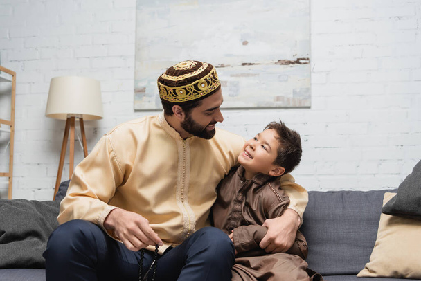 Hombre musulmán con cuentas de oración abrazando hijo en el sofá en casa  - Foto, Imagen