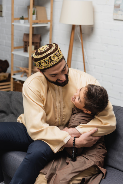 Alegre hombre musulmán con cuentas de oración abrazo hijo en el sofá en casa  - Foto, imagen