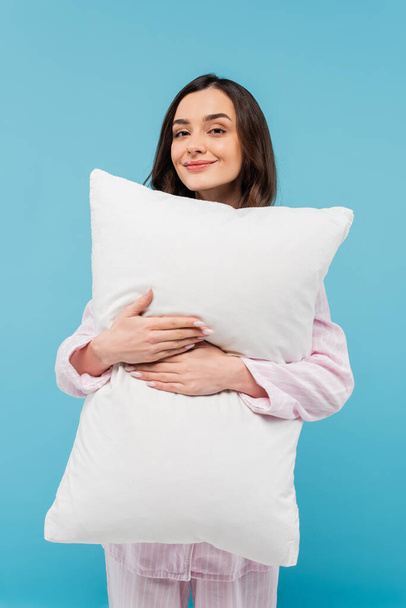 ευχαριστημένη νεαρή γυναίκα σε υπνόσακο κρατώντας λευκό μαξιλάρι απομονωμένο στο μπλε  - Φωτογραφία, εικόνα