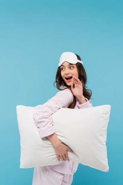 amazed young woman in nightwear and sleeping mask holding white pillow isolated on blue  - Fotoğraf, Görsel