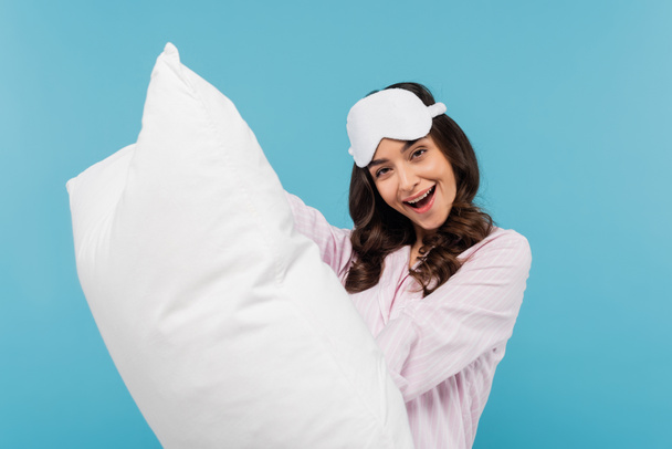femme excitée en vêtements de nuit et masque de sommeil tenant oreiller blanc isolé sur bleu  - Photo, image