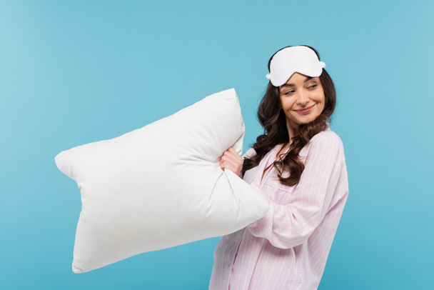 pleased young woman in pajamas and night mask holding white pillow isolated on blue  - Фото, изображение