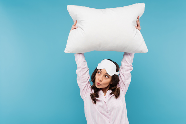 brunette young woman in pajamas and night mask holding white pillow while pouting lips isolated on blue  - Foto, afbeelding