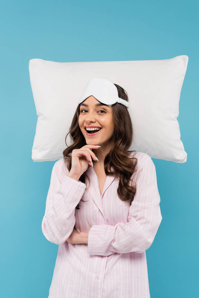 ενθουσιασμένοι νεαρή γυναίκα με πιτζάμες και μάσκα νύχτα χαμογελώντας κοντά λευκό ιπτάμενο μαξιλάρι απομονωμένο σε μπλε  - Φωτογραφία, εικόνα