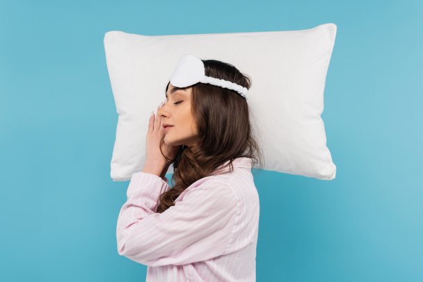 パジャマのブルネットの女性と青で隔離された白い枕の上で寝ている夜のマスクの側面図  - 写真・画像