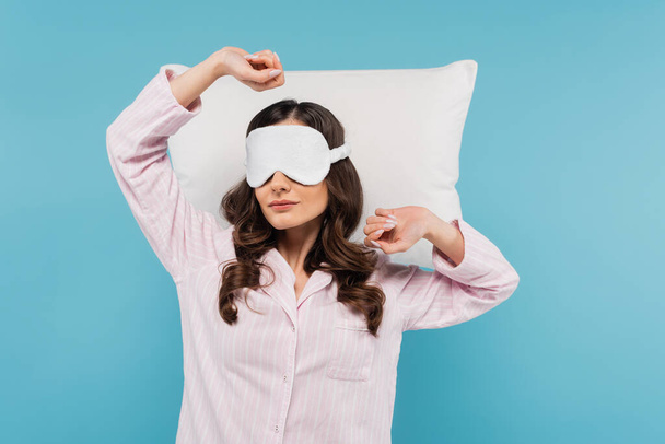 young woman in pajamas and night mask stretching near white pillow isolated on blue - Foto, Bild