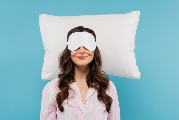 heureux jeune femme en pyjama et masque de nuit reposant sur oreiller blanc isolé sur bleu  - Photo, image